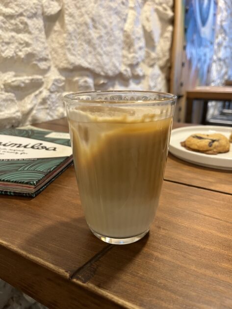 Un vaso de café helado en capas reposa sobre una mesa de madera, con un menú y un plato con una galleta en el fondo.
