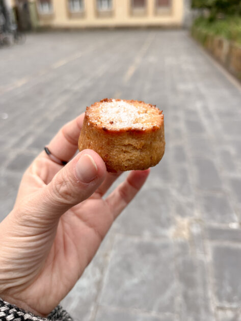 Una mano que sostiene un pastel pequeño y redondo espolvoreado con azúcar en polvo, con un fondo urbano exterior borroso.