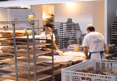 Dos panaderos dan forma a la masa sobre una mesa en la cocina de una panadería, rodeados de rejillas y bandejas de panes. En la camisa de uno de los panaderos se lee "Sourdough Bread & Baker.