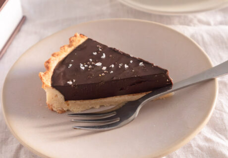 Una rebanada de tarta de chocolate con base de masa quebrada, cubierta con sal marina, servida en un plato con tenedor. Al fondo se ven dos rebanadas más.