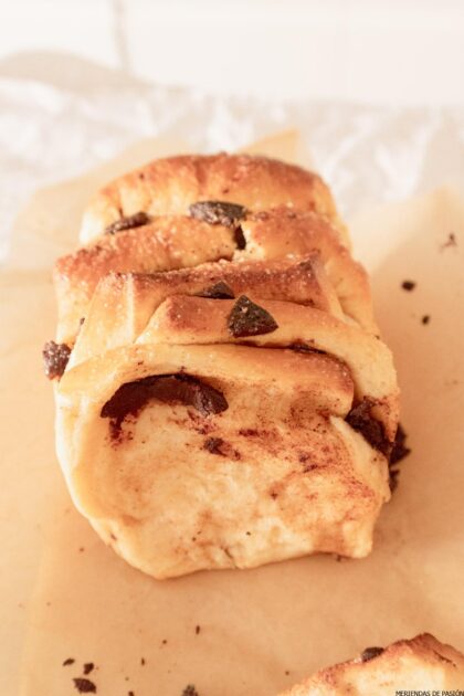 Una barra de pan para compartir con remolinos visibles de canela y trocitos de chocolate, colocada sobre papel de horno.