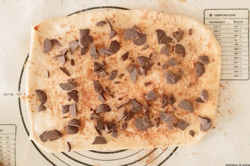 Masa extendida y espolvoreada con canela, azúcar y trocitos de chocolate sobre una esterilla de repostería con marcas de medida.